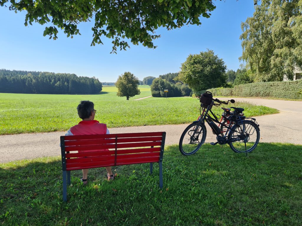 Person auf Bank, daneben Fahrräder, vor Blick auf hügeliges Gelände mit Solitärbaum, Wiesen und Wäldern